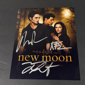 Puede incluir: Un póster promocional rectangular de The Twilight Saga: New Moon con tres actores en un bosque oscuro y una luna creciente. El póster incluye autógrafos en tinta plateada y el título de la película en letras amarillas y blancas.