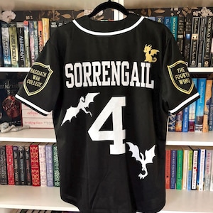 May include: A black baseball jersey featuring white text that reads SORRENGAIL above the number 4. The shirt includes white dragon silhouettes, a gold dragon icon, and two shield-shaped patches on the sleeves labeled Basgiath War College and The Fourth Wing.