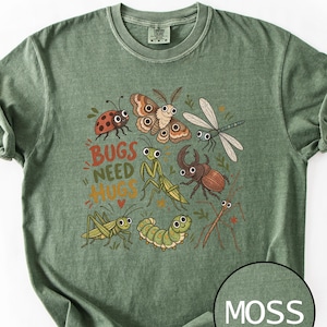May include: A moss green short-sleeve t-shirt featuring a graphic print of various cartoon insects including a ladybug, moth, dragonfly, beetle, mantis, caterpillar, and stick insect with the text "BUGS NEED HUGS" and a small red heart.