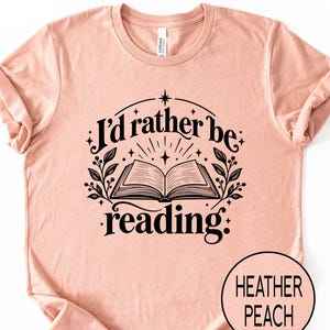 May include: A heather peach short-sleeve t-shirt featuring a black graphic of an open book with the text "I'd rather be reading" above and below the illustration, surrounded by decorative leaves and star accents.