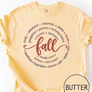 May include: A butter yellow t-shirt featuring a circular graphic design with the word fall in large cursive script at the centre. Surrounding the text are smaller words including sweaters, scarves, jeans, boots, apple cider, pie, autumn, hayrides, bonfires, turkey, candy corn, corn mazes, thanks, harvest, crunchy leaves, pumpkins, and patches.