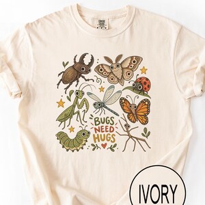 May include: An ivory short-sleeve t-shirt featuring a central graphic of various cartoon insects including a beetle, moth, ladybug, praying mantis, dragonfly, butterfly, caterpillar, and stick bug with the text "BUGS NEED HUGS" in brown letters.