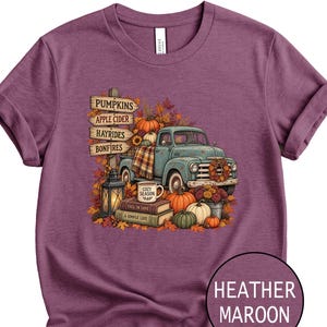 May include: A heather maroon t-shirt featuring a graphic of a teal vintage truck, pumpkins, a lantern, and books. The signpost reads PUMPKINS, APPLE CIDER, HAYRIDES, BONFIRES. A mug on the books says COZY SEASON, with text FALL IN LOVE and A SIMPLE LIFE.