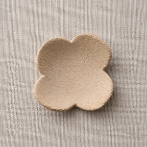 May include: A small, four-petaled flower-shaped dish with a textured, matte beige surface. The object features a shallow, concave centre and rounded edges, presenting a uniform, earthy tone throughout its entire visible structure.