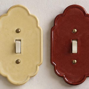May include: Two scalloped-edge light switch plates, one in a pale yellow and one in a deep reddish-brown, each featuring a central white toggle switch and two brass-colored screws positioned vertically above and below the switch mechanism.