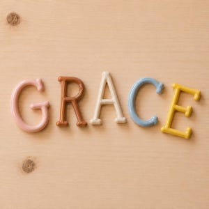 May include: Five individual letters spelling the word GRACE, each featuring a smooth, glossy finish. The letter G is light pink, R is terracotta brown, A is cream, C is dusty blue, and E is mustard yellow, all arranged in a horizontal line.