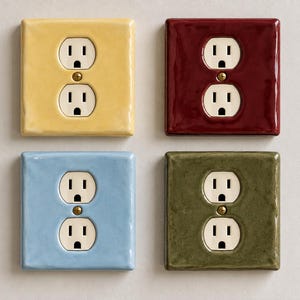 Pode incluir: Quatro espelhos de tomada quadrados com acabamento brilhante, dispostos em grade. As placas são amarelas, vermelho-escuras, azul-claras e verde-oliva, cada uma com uma tomada dupla branca e um parafuso central de latão.