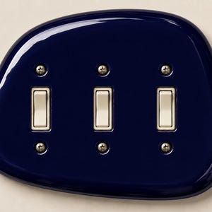 May include: A triple light switch wall plate with a glossy, dark navy blue finish and an irregular, rounded pebble-like shape. It features three cream-colored toggle switches arranged horizontally, each secured by two small silver-toned metal screws.