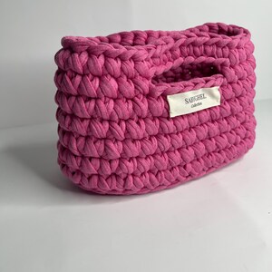Op de afbeelding: Een levendige fuchsia roze gehaakte mand met een dik, getextureerd breipatroon en geïntegreerde handvatten. Een rechthoekig gebroken wit stoffen label is op de voorkant genaaid, met de zwarte gedrukte tekst SABYGHEL Collection.