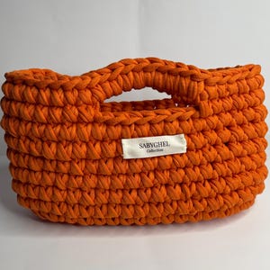 Op de afbeelding: Een feloranje, met de hand gehaakte mand met een dik, getextureerd weefsel en geïntegreerde handvatten. Een wit rechthoekig stoffen label is aan de voorzijde bevestigd, met de zwarte tekst SABYGHEL Collection in een duidelijk schreefloos lettertype.