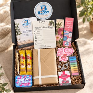 May include: A black gift box filled with brown paper crinkle fill containing a wrapped book, two Nescafé coffee sticks, a pen, a tin of Big Sky Mints, a Reader's Date Card, two bookmarks, a set of colorful sticky tabs, and decorative stickers.
