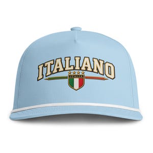 May include: A light blue baseball cap featuring a white cord across the brim. The front displays the word ITALIANO in arched white letters with a black outline, positioned above a shield emblem with the Italian flag and four gold stars.