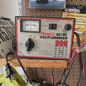 May include: A Hobbico AC/DC Multi-Charger 905 with a silver faceplate, red border, and black control knob. The device features an analog ammeter, a discharge/charge toggle switch, and a 1-amp fuse holder. Text includes DC Input 12 volts, AC Input 110 volts.