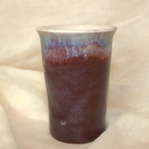 May include: A cylindrical ceramic tumbler featuring a deep reddish-brown base glaze that transitions into a lighter, mottled blue and white glaze near the flared rim. The surface has a smooth, glossy finish with subtle colour variations throughout.