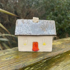 May include: A small ceramic house figurine with a smooth off-white exterior, a textured dark grey roof, and a central chimney. The front facade features a bright orange rectangular door and two small windows with yellow triangular accents.