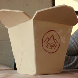 May include: A cream-colored ceramic container shaped like a traditional takeout box with folded top flaps. The front features a hand-drawn red circular emblem containing a mountain graphic and the handwritten text "Enjoy" in red ink.