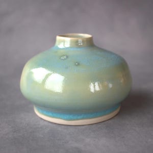 May include: A small, round ceramic vase with a glossy finish. The glaze features a blend of soft sage green and light blue tones, with a darker blue band near the base. The vase has a short, narrow neck and a smooth, rounded body shape.