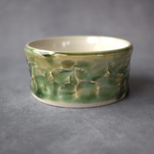 May include: A small, round ceramic bowl featuring a textured, dimpled exterior surface with a glossy glaze in shades of sage green, olive, and cream. The interior of the bowl is smooth and finished in a solid, off-white cream color.