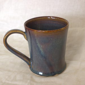 May include: A ceramic mug featuring a glossy glaze with blended tones of deep brown, dark blue, and hints of amber. The mug has a cylindrical body with a slightly flared base and a curved handle attached to the side, showcasing a smooth finish.