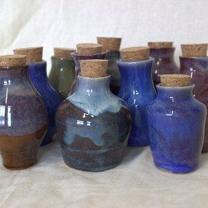 May include: A collection of small, handmade ceramic jars with cork stoppers. Each jar features a unique, glossy glaze in various shades of blue, purple, and brown, with some showing blended color transitions. The jars have a rounded body and a narrow neck.