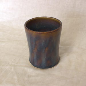 May include: A cylindrical ceramic tumbler featuring a glossy glaze with blended tones of deep brown, amber, and dark blue. The vessel has a smooth, slightly tapered body with a wide circular opening and a solid base, showcasing a reactive finish.
