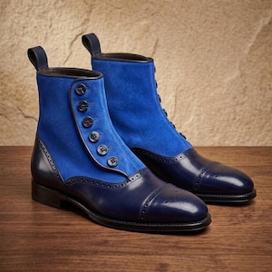 Op de afbeelding: Een paar enkellaarzen met een bovenwerk van felblauw suède en een donkerblauwe leren neus en hiel. De laarzen hebben een zijdelingse knoopsluiting met zeven donkere knopen en decoratieve brogue-details langs de leren randen.