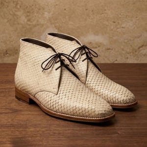 May include: A pair of ankle boots featuring a textured, cream-colored scale pattern across the exterior. The boots have a rounded toe, a low heel, and dark brown laces threaded through eyelets. The construction includes a visible brown sole edge.