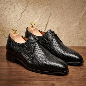 May include: A pair of black lace-up dress shoes featuring a textured snakeskin-like pattern on the leather upper. Each shoe includes a light-colored wooden shoe tree inserted inside to maintain the shape of the footwear.