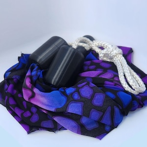 May include: A jump rope featuring two solid black cylindrical handles and a white braided rope. The rope is coiled atop a piece of fabric patterned with a black, blue, and purple mosaic design that resembles a giraffe or stone texture.