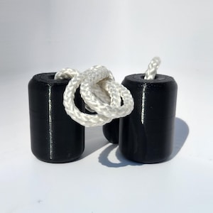May include: Two cylindrical black weights with rounded edges, each featuring a central hole. A thick, white braided rope is threaded through the holes of both cylinders, connecting them and forming a loose loop between the two black components.