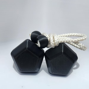 Peut inclure: Deux objets noirs de forme pentagonale avec une finition mate, reliés par un cordon tressé blanc. Chaque objet présente une face avant plate et lisse et est fixé au cordon par une petite perle sphérique noire, créant un design géométrique.