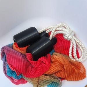 May include: A pair of black cylindrical handles attached to a white braided rope, resting on a pile of pleated, multi-colored fabric in shades of red, orange, blue, and purple. The fabric has a textured, crinkled appearance throughout the material.