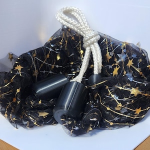 May include: A black sheer fabric pouch featuring a gold and silver star pattern, cinched with a thick white braided rope drawstring. The closure is secured by two smooth, cylindrical black plastic beads attached to the white cord ends.