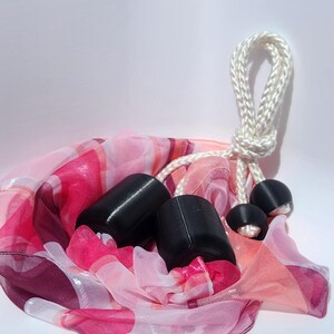 May include: A sheer scarf featuring a vibrant pattern of pink, magenta, and white stripes and circles. The fabric is gathered and secured by a white braided rope cord that features two large black cylindrical weights and two smaller black spherical beads.