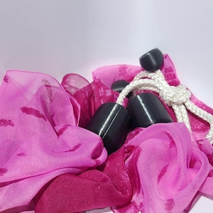 May include: A vibrant pink sheer fabric with a subtle darker pink pattern, featuring several smooth black cylindrical beads attached to a thick white braided cord. The fabric is draped in soft folds, contrasting with the solid black beads.