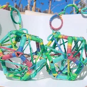 May include: Two geometric bird toys constructed from interconnected pentagonal frames wrapped in multicolored yarn in shades of green, blue, yellow, and pink. Each toy features hanging rings and a smooth white spherical bead at the end.