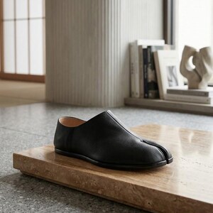 May include: A black leather slip-on shoe featuring a distinct split-toe design, a smooth rounded silhouette, a low profile, and a thin sole. The shoe has a visible center seam running from the toe split to the opening, with a tan interior lining.