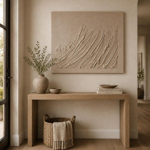 May include: A rectangular beige textured wall art piece with raised diagonal strokes hangs above a light wood console table. On the table sits a ceramic vase with green stems, a small bowl, and two stacked books labeled Picasso and Kinfolk.