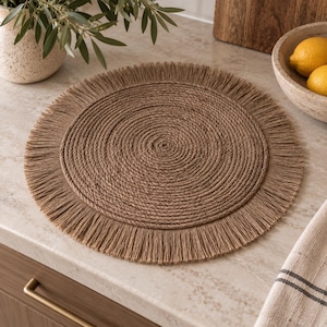 May include: A round placemat constructed from coiled brown rope, featuring a textured center and a decorative fringed border. The circular design is composed of tightly wound natural-toned fibers, creating a consistent pattern throughout the surface.