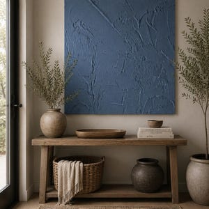 May include: A square wall art piece with a deep blue, heavily textured impasto surface. The artwork hangs above a wooden console table featuring a ceramic vase, a wooden bowl, a stack of books, a woven basket with a throw, and a dark pot.