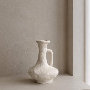 Może przedstawiać: Kremowy wazon ceramiczny o teksturowanej, nierównej powierzchni. Naczynie posiada szeroką, zaokrągloną podstawę, która zwęża się w smukłą szyjkę, zakończoną rozszerzonym brzegiem. Pojedynczy, zakrzywiony uchwyt jest przymocowany do boku szyjki.