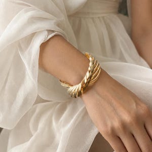May include: A gold-toned cuff bracelet featuring a twisted, multi-layered design with a textured, brushed metal finish. The bracelet is open-ended and wraps around the wrist, showcasing a series of parallel, curved metal bands that create a dimensional, spiraling effect.