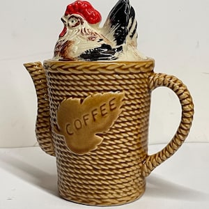 May include: A ceramic coffee pot with a brown, rope-textured exterior and a leaf-shaped emblem embossed with the word COFFEE. The lid features a sculpted, hand-painted chicken with a bright red comb, white feathers, and black tail accents.