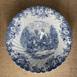 May include: A round ceramic bowl with a scalloped edge featuring a blue and white transferware pattern. The center depicts a horse-drawn carriage scene, while the border displays floral motifs. A small white sticker with the number 30 is visible.