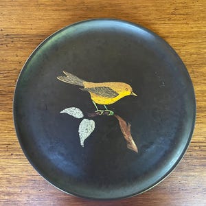 May include: A round, matte black plate featuring a central illustration of a yellow bird perched on a brown branch with two textured, light-colored leaves. The bird has detailed wing markings and a small beak, centered on the dark surface.