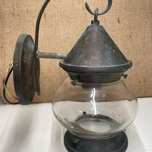 May include: A wall-mounted lantern featuring a weathered copper finish with a conical top, a curved support arm, and a clear, rounded glass globe. The fixture includes a circular mounting plate and exposed electrical wiring at the back.
