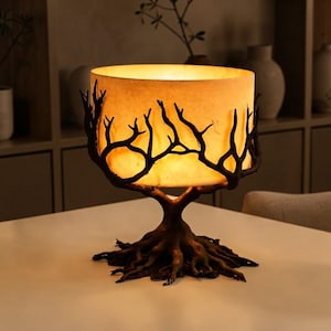 May include: A table lamp featuring a cylindrical, warm-glowing shade surrounded by a dark, textured base shaped like tree roots and branches that extend upward to cradle the light. The base has a weathered, organic appearance with intricate details.