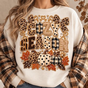 May include: A cream-coloured crewneck sweatshirt featuring a graphic print with the text COZY SEASON. The design includes leopard print, buffalo plaid, and polka dot patterns, two large bows, three pumpkins, and autumn leaves with rhinestone accents.