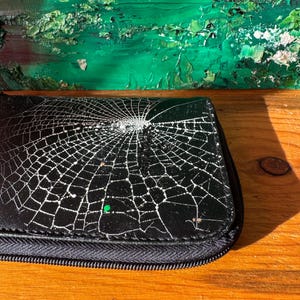 May include: A black rectangular wallet featuring a white spiderweb graphic printed across the surface. The wallet has a black zipper closure along the edge and a single small green dot detail integrated into the web pattern design.