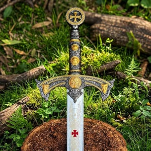 Könnte beinhalten: Ein dekoratives Schwert mit einem verzierten silber-goldenen Griff, kreisförmigen Emblemen und einem Kreuzknauf. Das Parierstange zeigt komplizierte Schnörkel und goldene Schilde. Die Klinge ist mit filigranen Gravuren und einem roten Kreuz versehen.